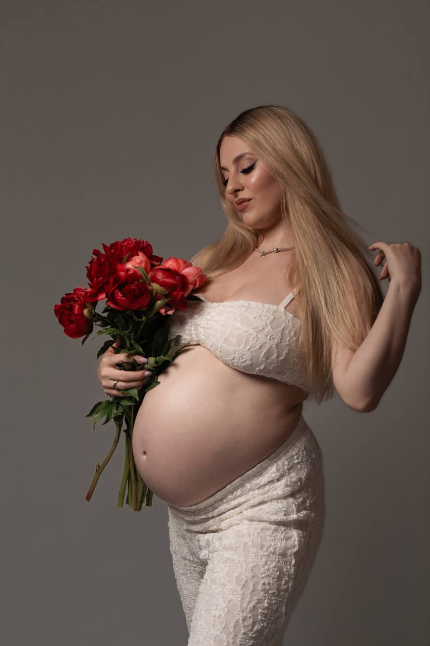 Expecting mother in white lace holding a bouquet of red peonies