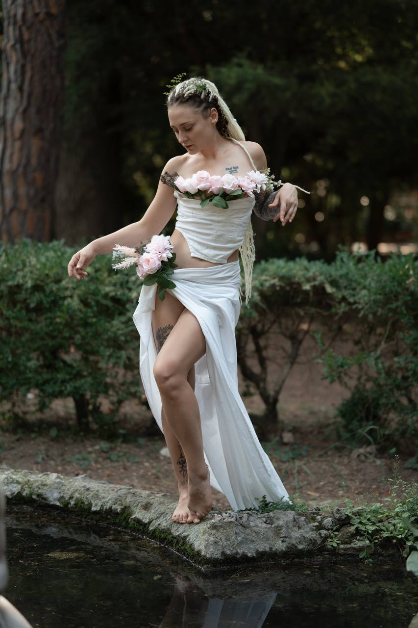 A woman in flowing white with a flower crown standing at the edge of a pond