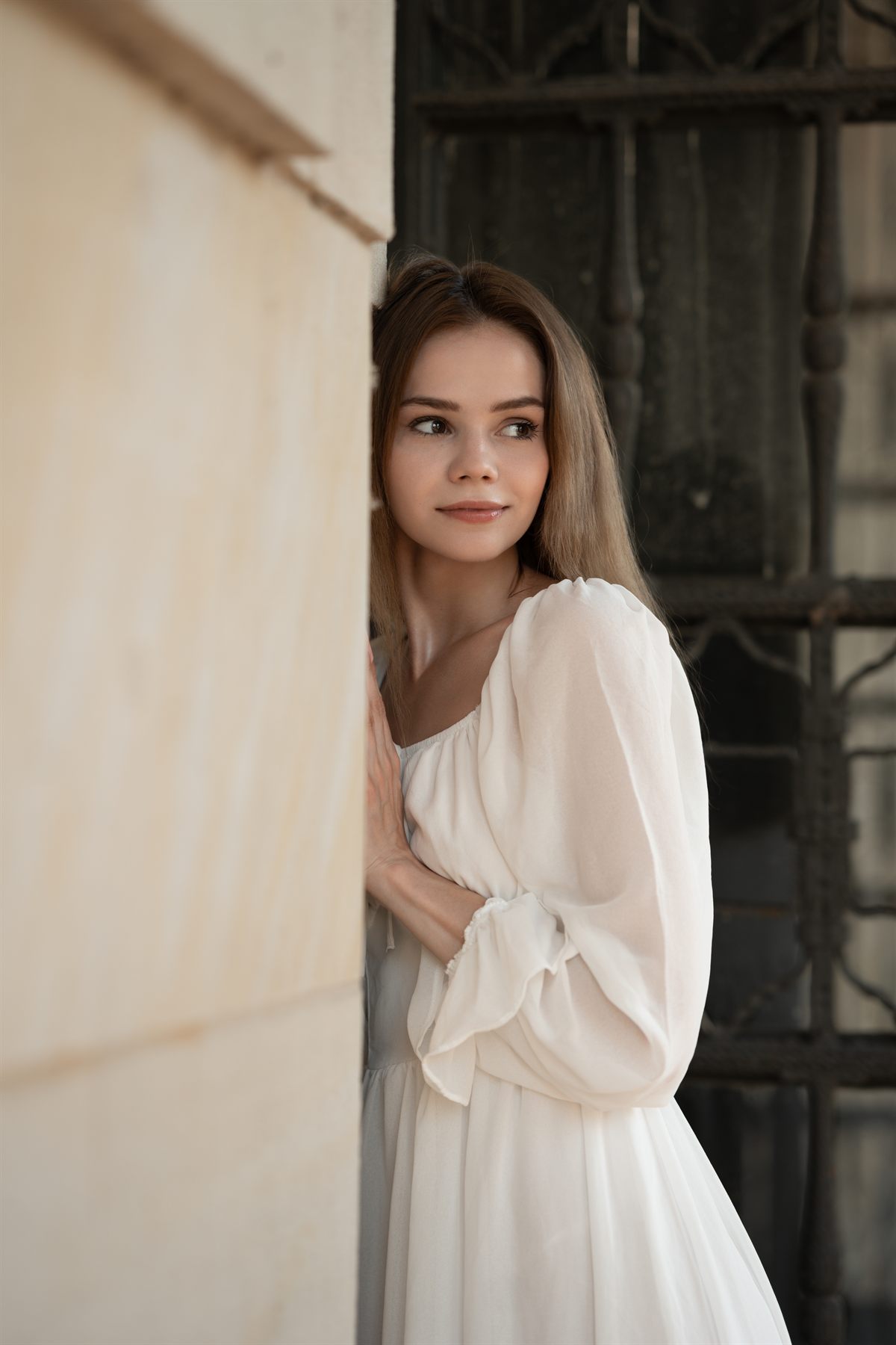 A young woman in a soft white dress leaning against a sunlit wall