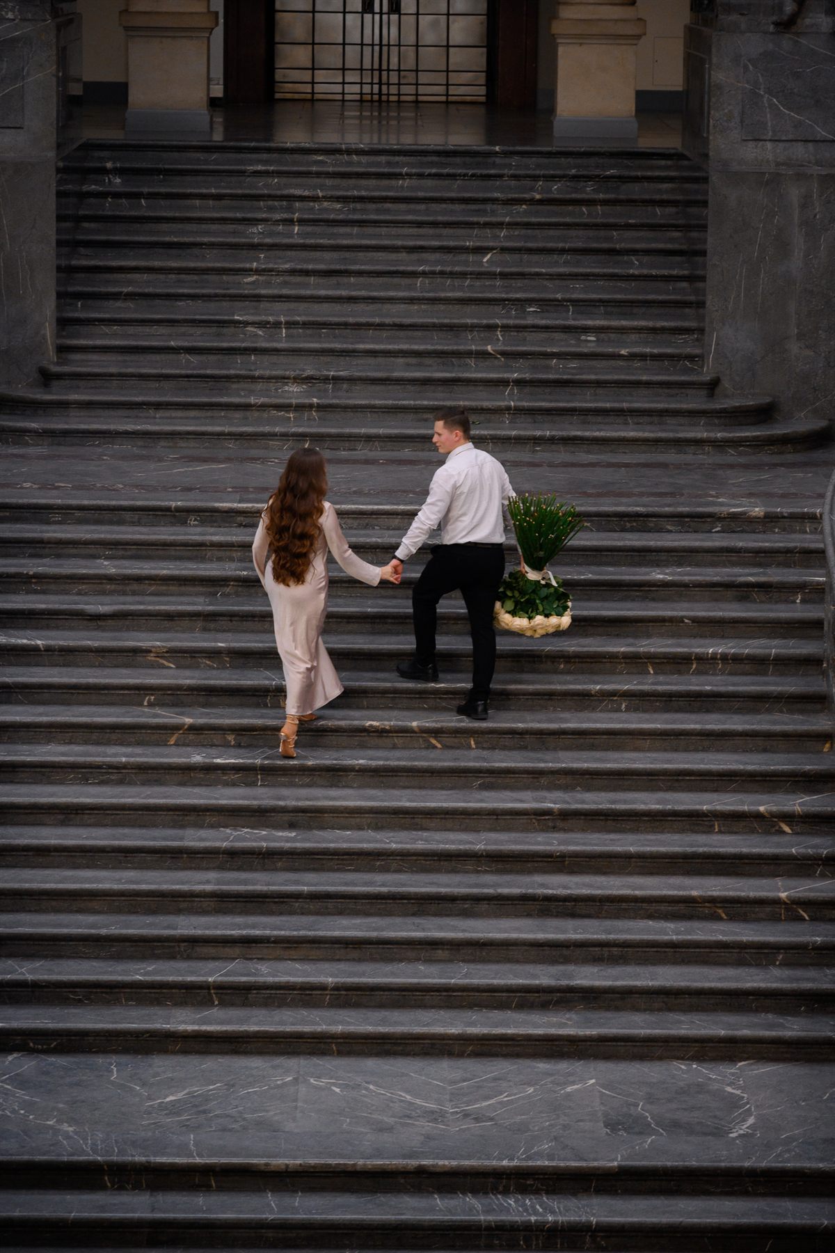 A couple climbing a wide marble staircase, carrying a bouquet of white roses