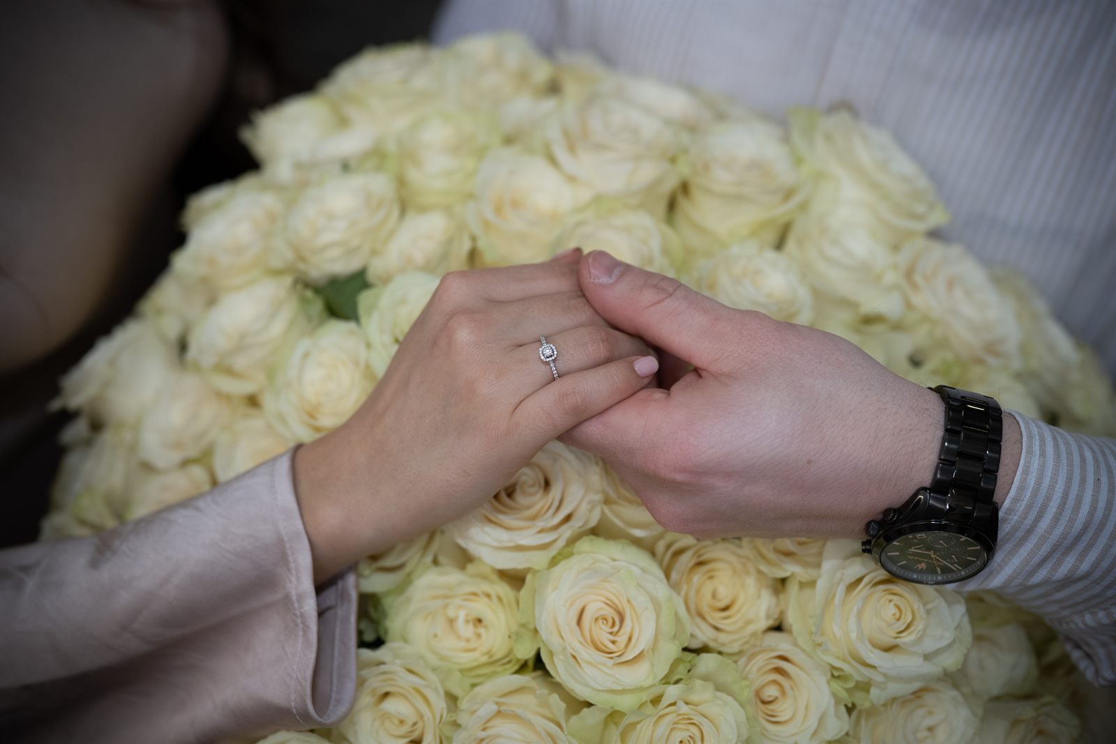 Her hand with the engagement ring resting on his, above white roses