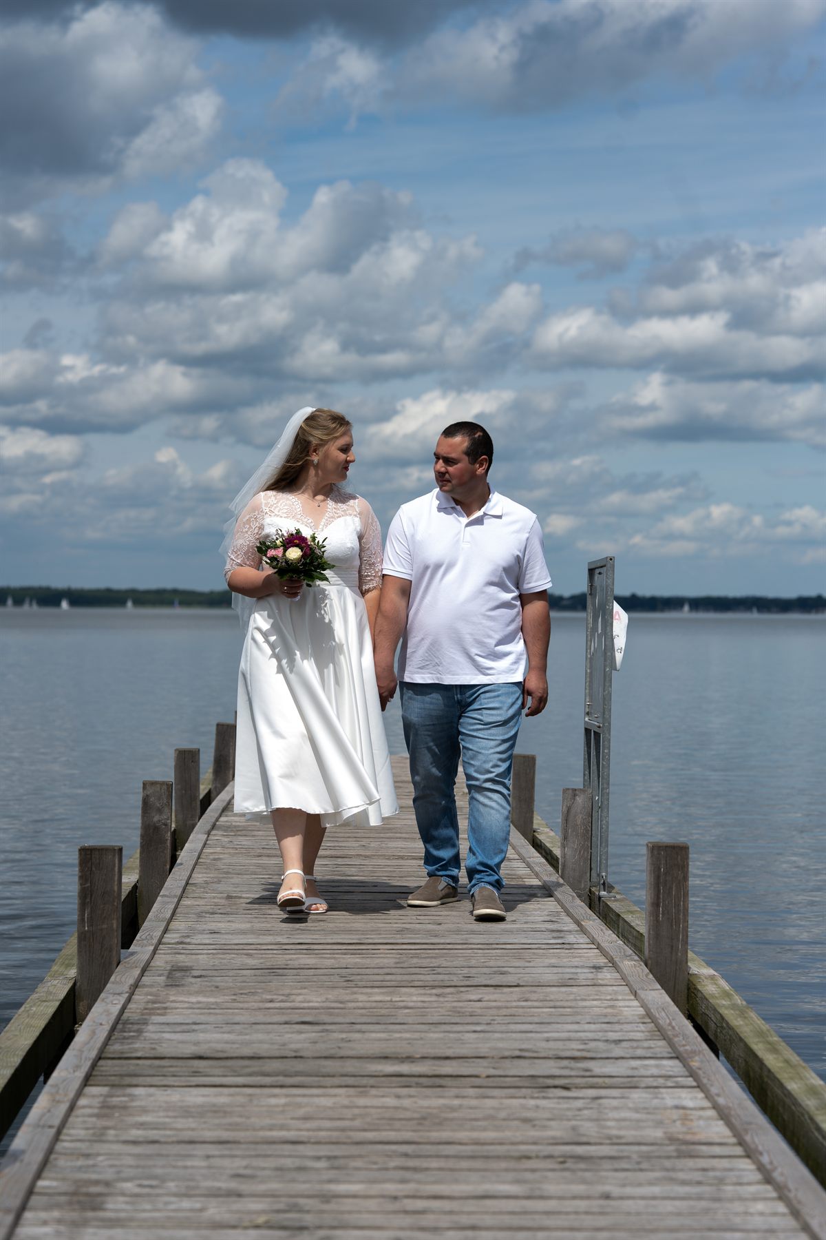 A bride and groom walking hand in hand along a wooden pier over the water