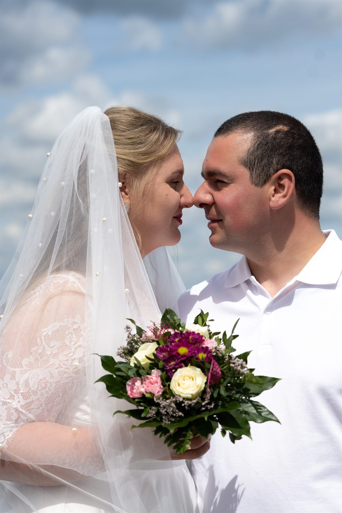 A bride and groom nose to nose against a bright summer sky